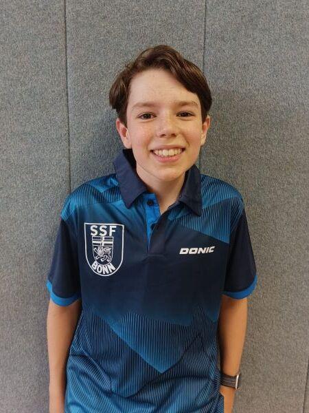 Junger Sportler mit fr&ouml;hlichem L&auml;cheln tr&auml;gt ein blaues Trikot mit einem Emblem und steht vor einer grauen Wand.