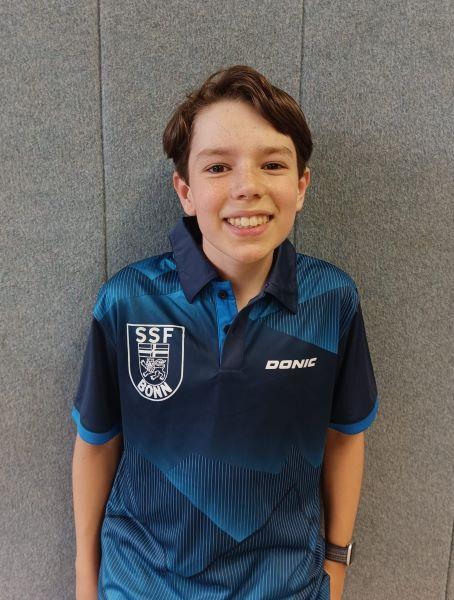 Junger Sportler mit fröhlichem Lächeln trägt ein blaues Trikot mit einem Emblem und steht vor einer grauen Wand.
