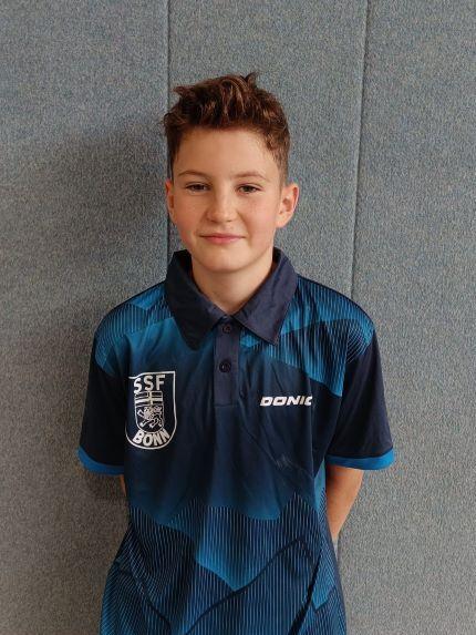 Portr&auml;t eines Jungen in einem blauen Trikot mit dem Logo des SSF Bonn, vor einer grauen Wand.