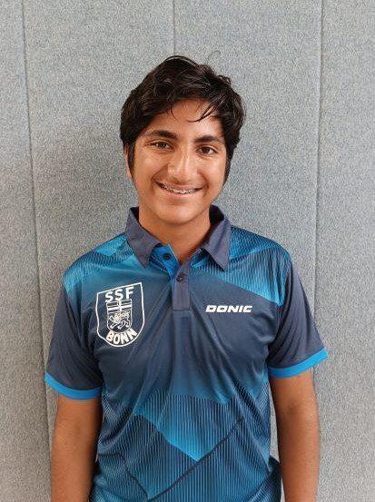 Junge mit dunklen Haaren tr&auml;gt ein blaues Sportshirt mit Logo und l&auml;chelt vor einer grauen Wand.