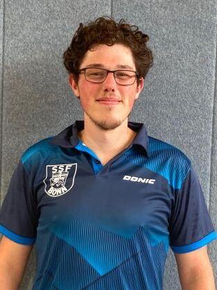 Junger Mann in blauer Sportbekleidung steht vor einer grauen Wand und l&auml;chelt in die Kamera.