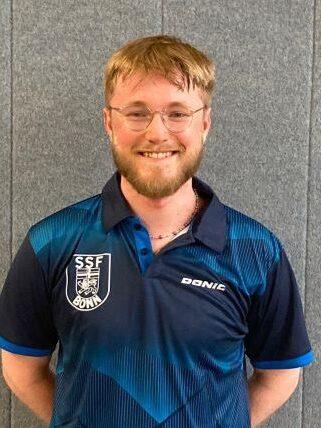 L&auml;chelnder junger Mann mit Brille in einem blauen Sporttrikot vor einer grauen Wand.