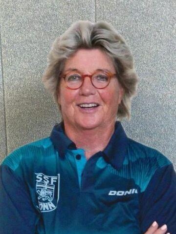 L&auml;chelnde Frau mit kurzen grauen Haaren in sportlichem Polohemd, mit verschr&auml;nkten Armen vor einer hellen Wand.