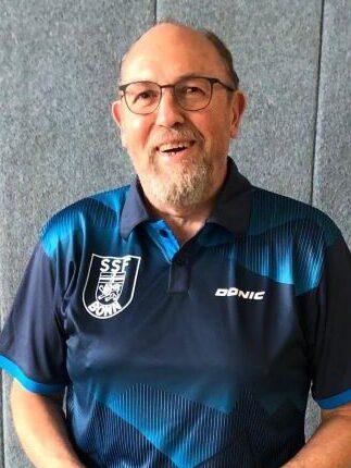&Auml;lterer Mann mit Brille steht l&auml;chelnd vor einer grauen Wand, tr&auml;gt ein blaues Tischtennisshirt mit Vereinslogo.