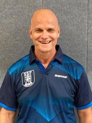 Mann mit Glatze tr&auml;gt ein blaues Tischtennis-Trikot und l&auml;chelt vor einer grauen Wand.