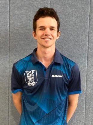 Ein junger Mann steht l&auml;chelnd vor einer grauen Wand, tr&auml;gt ein blaues Sporttrikot mit dem Logo &bdquo;SSF Bonn&ldquo;.