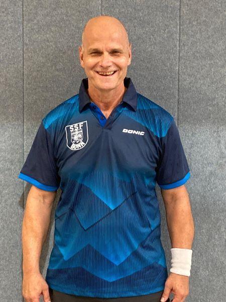 Mann mit Glatze tr&auml;gt ein blaues Tischtennis-Trikot und l&auml;chelt vor einer grauen Wand.