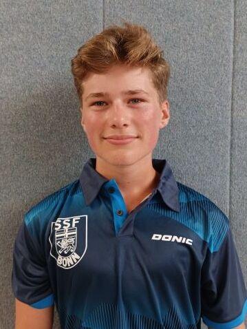 Portr&auml;t eines jungen Mannes in einem blauen Tischtennis-Trikot mit Aufdruck, l&auml;chelnd vor grauem Hintergrund.