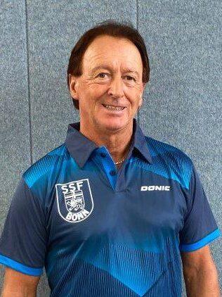 Mann in einem blauen Tischtennisshirt mit Logo steht vor einer grauen Wand und l&auml;chelt.