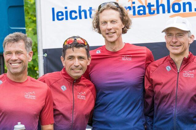 Vier Sportler in roten und blauen Trikots posieren l&auml;chelnd vor einem Banner mit dem Schriftzug &bdquo;leicht triathlon&ldquo;.