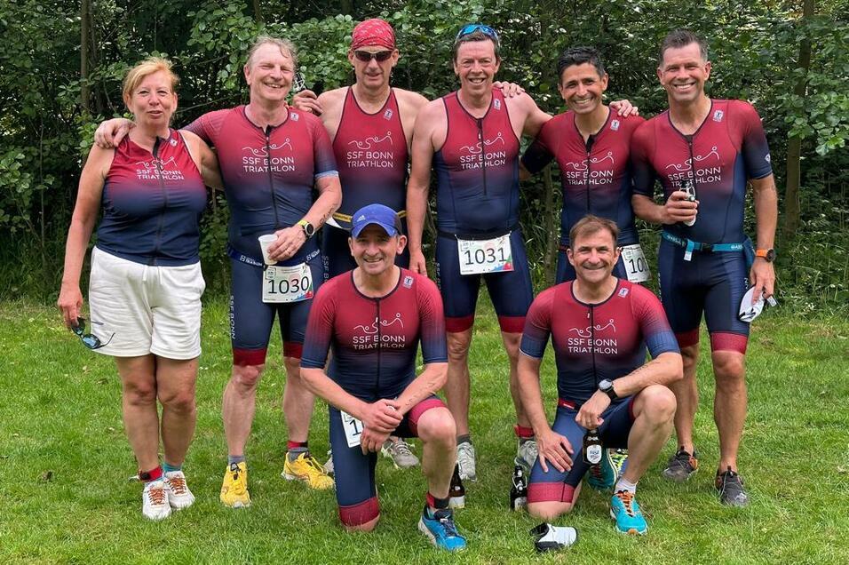 Gruppe von acht Triathleten in Sportkleidung posiert l&auml;chelnd auf einer Wiese nach einem Wettkampf.