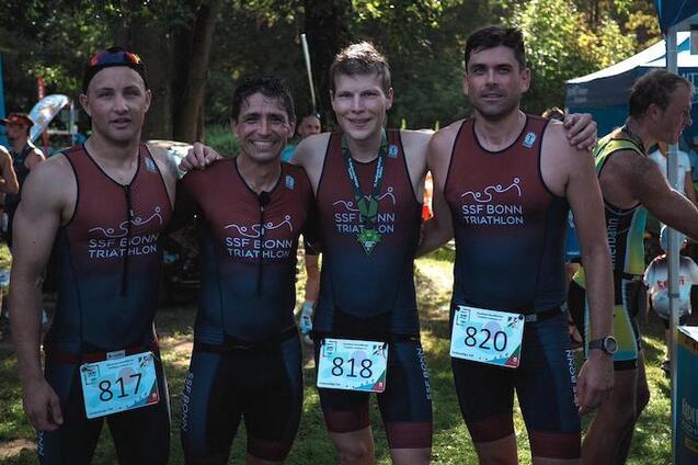Vier Triathleten des SSF Bonn stehen l&auml;chelnd zusammen, tragen Wettkampfanz&uuml;ge und haben Startnummern sichtbar.