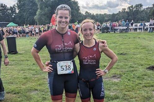 Zwei Triathletinnen in Sportanz&uuml;gen mit "SSF Bonn Triathlon"-Aufdruck posieren l&auml;chelnd vor einer Wettkampfszene.