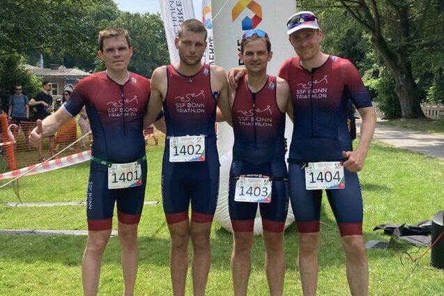 Vier m&auml;nnliche Triathleten in Sportkleidung stehen l&auml;chelnd auf einer Wiese vor einem Wettkampf-Startbogen.