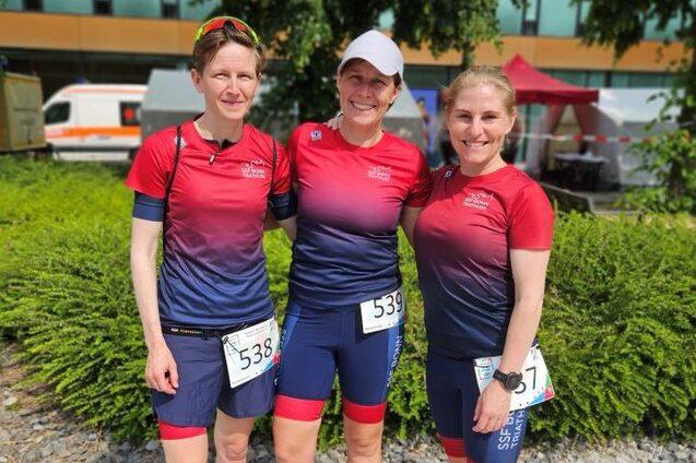 Drei Frauen in Sportkleidung mit Nummern auf einer Veranstaltung im Freien, umgeben von gr&uuml;nen B&uuml;schen und Zelt.