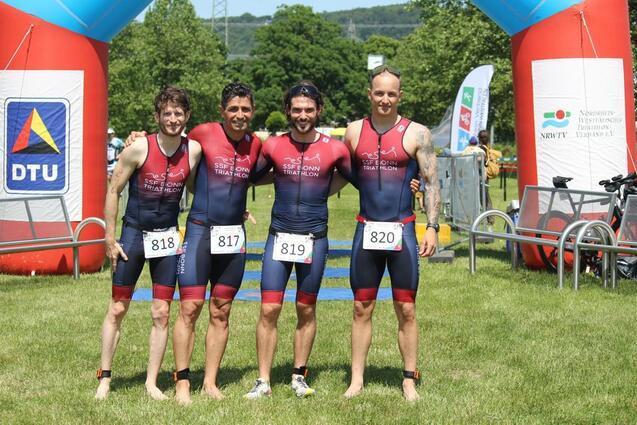 Vier Triathleten in einheitlichen Anz&uuml;gen stehen l&auml;chelnd vor einem Tor aus Luftballons bei einer Wettkampfveranstaltung.