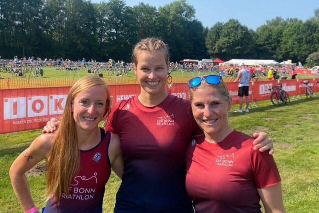 Drei Frauen in Sportkleidung l&auml;cheln vor einer Triathlon-Veranstaltung mit Zuschauern und Zelt im Hintergrund.