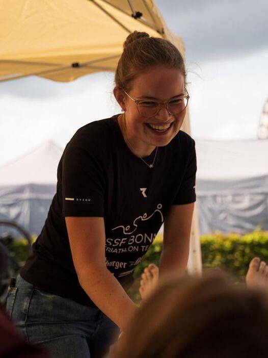 Eine l&auml;chelnde Frau in einem T-Shirt steht unter einem Zelt und gibt einer Person eine Massage.