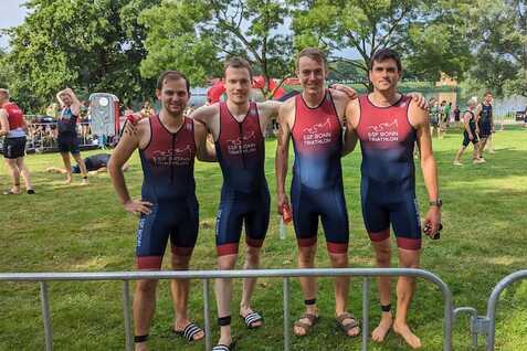 Vier Athleten in einheitlichen Triathlonanz&uuml;gen posieren auf einer gr&uuml;nen Wiese am Veranstaltungsort eines Triathlons.