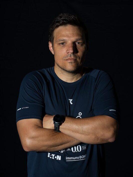 Ein sportlicher Mann mit kurzen Haaren steht mit verschr&auml;nkten Armen vor einem schwarzen Hintergrund.