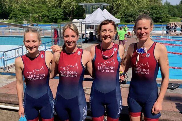 Vier Triathletinnen in einheitlichen Anz&uuml;gen posieren l&auml;chelnd vor einem Schwimmbecken unter blauem Himmel.