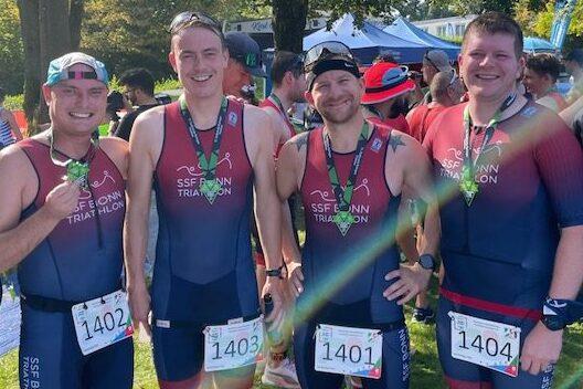 Vier M&auml;nner in Triathlon-Anz&uuml;gen stehen l&auml;chelnd zusammen, jeweils mit Medaillen und Startnummern an einem Wettkampf.