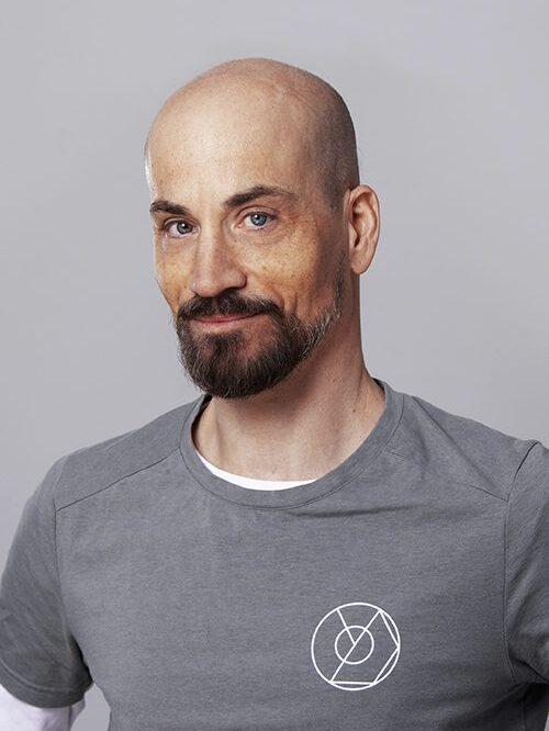 Mann mit Vollem Bart und Glatze l&auml;chelt, tr&auml;gt ein graues T-Shirt mit wei&szlig;em Logo, vor grauem Hintergrund.