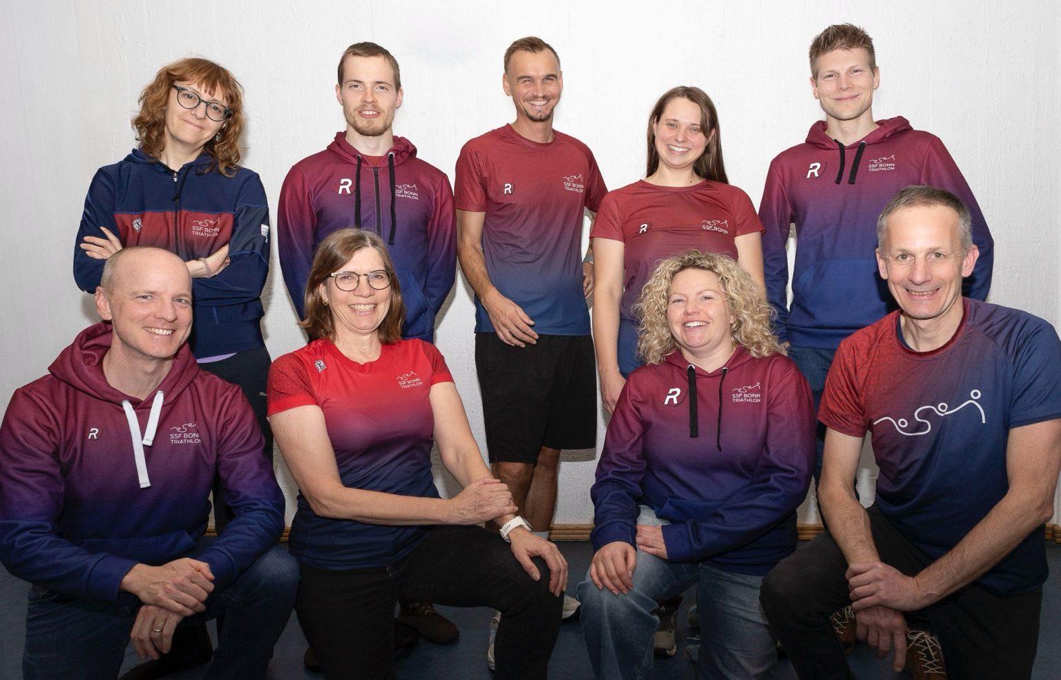 Gruppe von neun Personen in sportlichen Oberteilen, stehend und sitzend, l&auml;cheln vor neutralem Hintergrund.