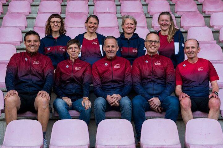 Gruppenshot von acht Personen in Sportkleidung, sitzend auf pinken Stadionb&auml;nken, l&auml;chelnd in die Kamera.