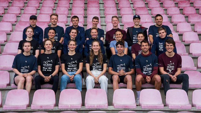 Gruppenfoto von 20 Personen in Teambekleidung, sitzend auf einer pinken Trib&uuml;ne in einem Stadion.