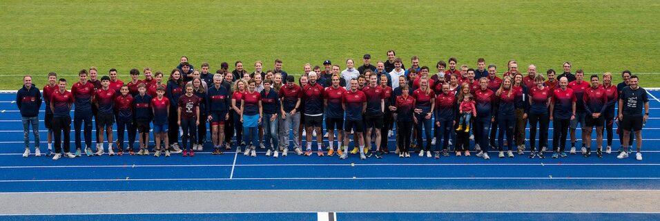 Gruppenfoto von Sportlern in roten Shirts auf einer blauen Laufbahn im Freien, umgeben von gr&uuml;nem Rasen.