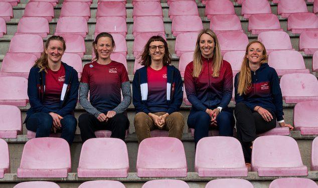 F&uuml;nf Personen in Sportkleidung sitzen l&auml;chelnd auf rosa Stadionpl&auml;tzen.