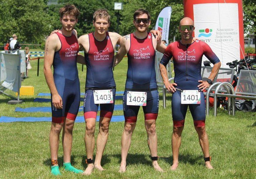 Vier Triathleten in Wettkampfanz&uuml;gen stehen l&auml;chelnd auf einer Wiese, bereit f&uuml;r den Wettkampf.