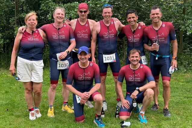 Gruppe von acht Triathleten in Sportkleidung posiert l&auml;chelnd auf einer Wiese nach einem Wettkampf.