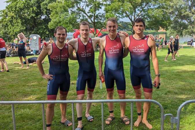 Vier Athleten in einheitlichen Triathlonanz&uuml;gen posieren auf einer gr&uuml;nen Wiese am Veranstaltungsort eines Triathlons.