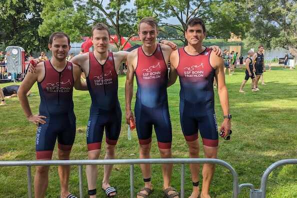 Vier Athleten in einheitlichen Triathlonanz&uuml;gen posieren auf einer gr&uuml;nen Wiese am Veranstaltungsort eines Triathlons.