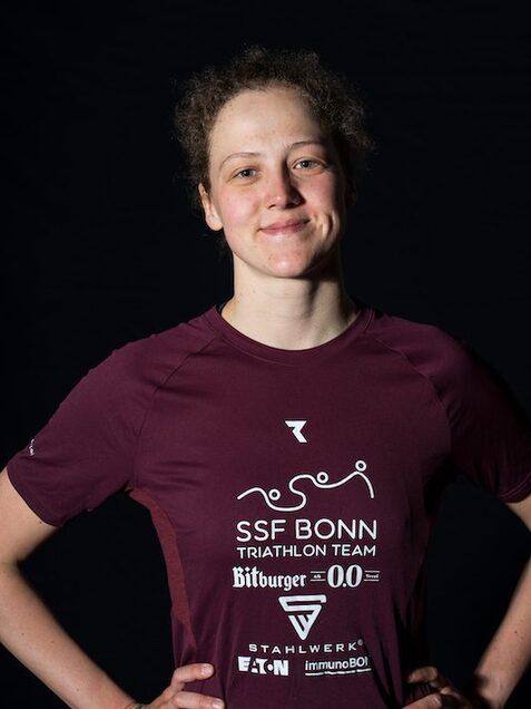 Portr&auml;t einer jungen Athletin in einem roten Triathlon-Shirt mit dem Aufdruck &bdquo;SSF Bonn Triathlon Team&ldquo; auf dunklem Hintergrund.
