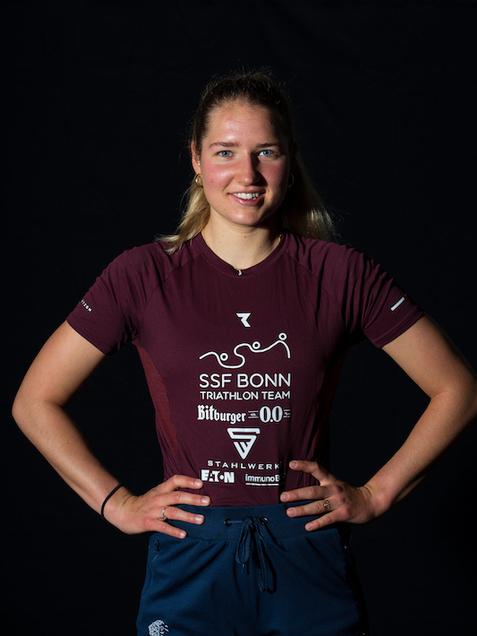 Portr&auml;t einer Sportlerin im roten Triathlon-Trikot des SSF Bonn, l&auml;chelnd mit H&auml;nden in der H&uuml;fte vor schwarzem Hintergrund.