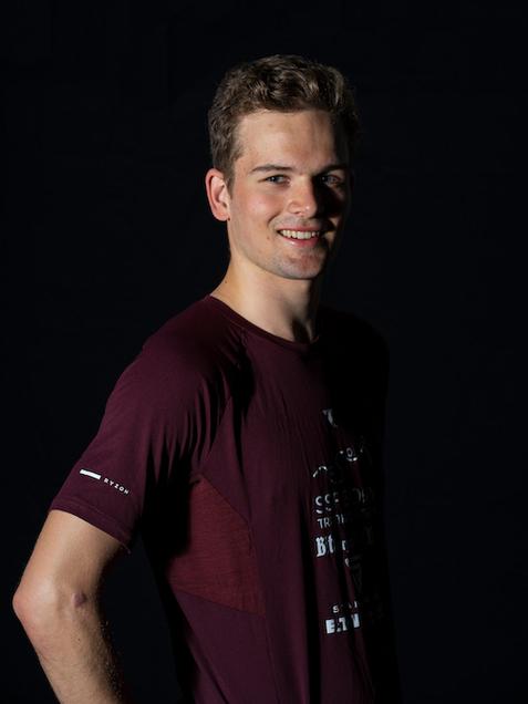 Portr&auml;t eines l&auml;chelnden jungen Mannes in einem dunkelroten T-Shirt vor schwarzem Hintergrund.