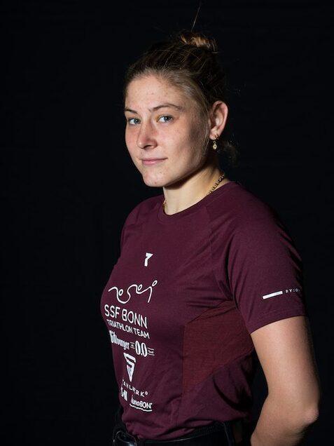 Junge Athletin in einem maroonen Triathlon-Shirt, seitlicher Blick vor schwarzem Hintergrund.