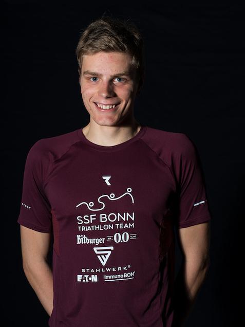 L&auml;chelnder junger Mann in einem burgunderfarbenen Triathlonsportshirt vor schwarzem Hintergrund.