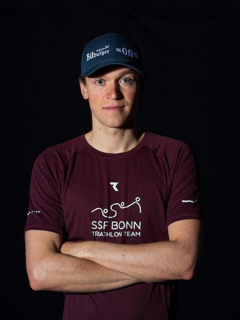 Portrait eines Mannes mit verschr&auml;nkten Armen, tr&auml;gt ein rotes T-Shirt des SSF Bonn Triathlon Teams und eine blaue Cap.