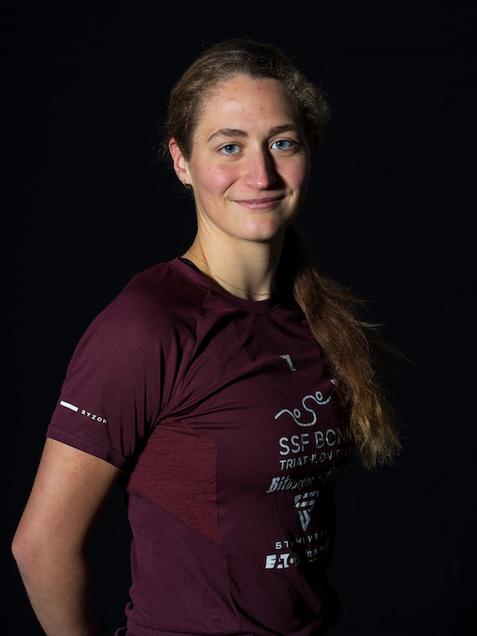 Portrait einer sportlichen Frau mit langen, lockigen Haaren, die in einem maroonfarbenen Triathlon-Trikot l&auml;chelt.