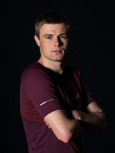 Portr&auml;t eines m&auml;nnlichen Sportlers mit kurzen Haaren, verschr&auml;nkten Armen und einem burgunderfarbenen T-Shirt vor dunklem Hintergrund.