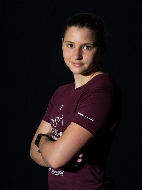 Junge Frau mit verschr&auml;nkten Armen, tr&auml;gt ein rotes T-Shirt, vor schwarzem Hintergrund. Blick in die Kamera.