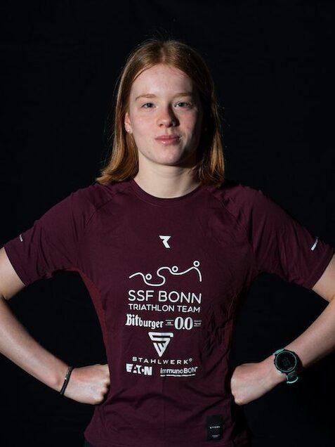 Portr&auml;t einer jungen Frau in einem burgunderfarbenen Triathlon-Shirt, mit H&auml;nden auf der H&uuml;fte vor schwarzem Hintergrund.