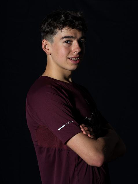 Portr&auml;t eines jungen Mannes mit Brille und T-Shirt, l&auml;chelnd und mit verschr&auml;nkten Armen vor dunklem Hintergrund.