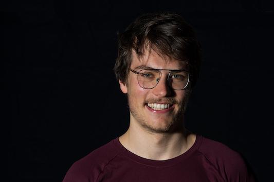 L&auml;chelnder Mann mit Brille tr&auml;gt ein rotes T-Shirt des SSF Bonn Triathlon Teams vor schwarzem Hintergrund.
