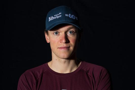 Portrait eines Mannes mit verschr&auml;nkten Armen, tr&auml;gt ein rotes T-Shirt des SSF Bonn Triathlon Teams und eine blaue Cap.