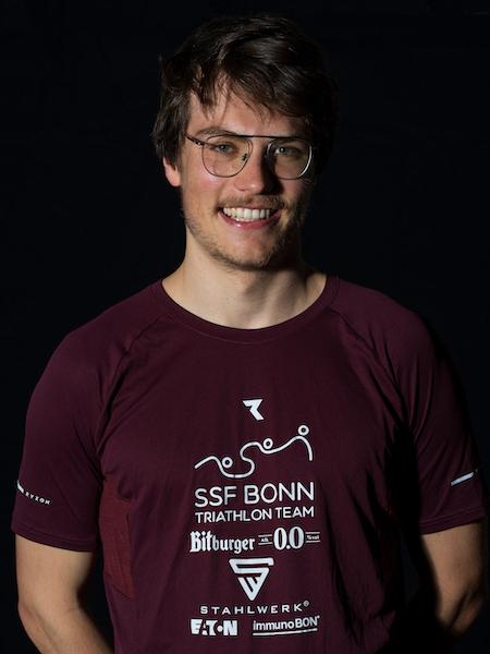 L&auml;chelnder Mann mit Brille tr&auml;gt ein rotes T-Shirt des SSF Bonn Triathlon Teams vor schwarzem Hintergrund.